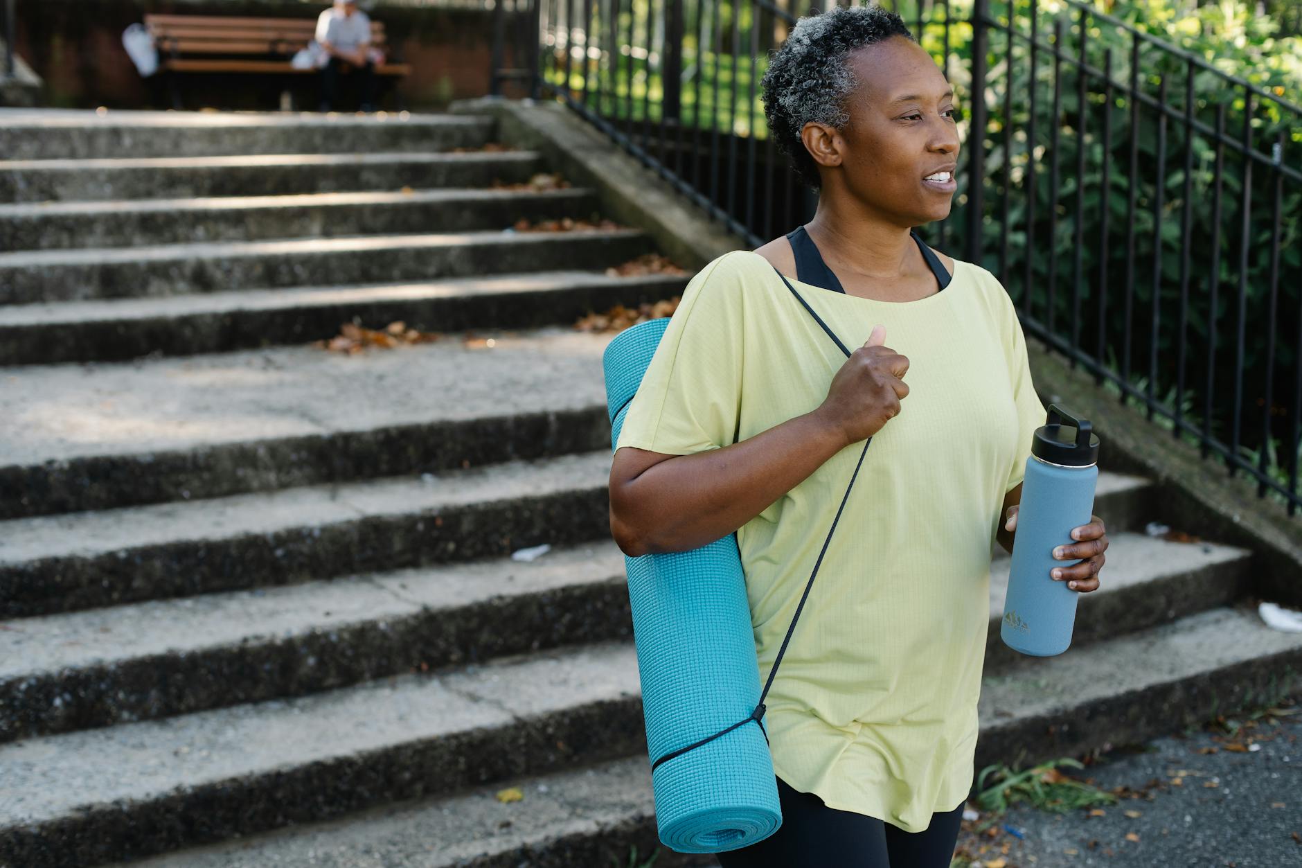 a woman in yellow shirt carrying yoga mat and tumbler